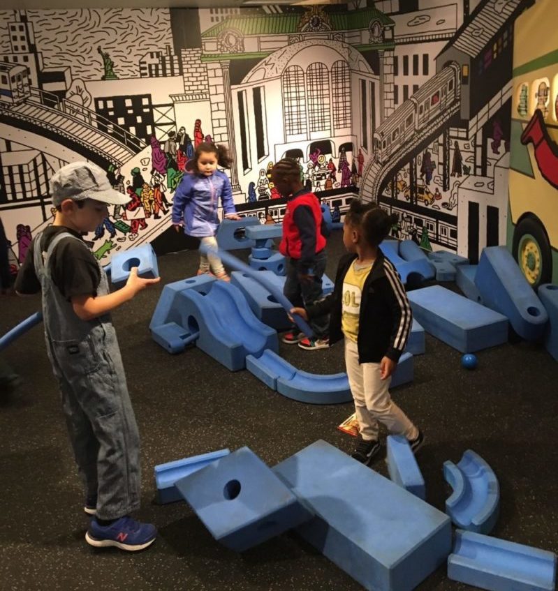 Four children stand or walk among big blue foam play blocks.