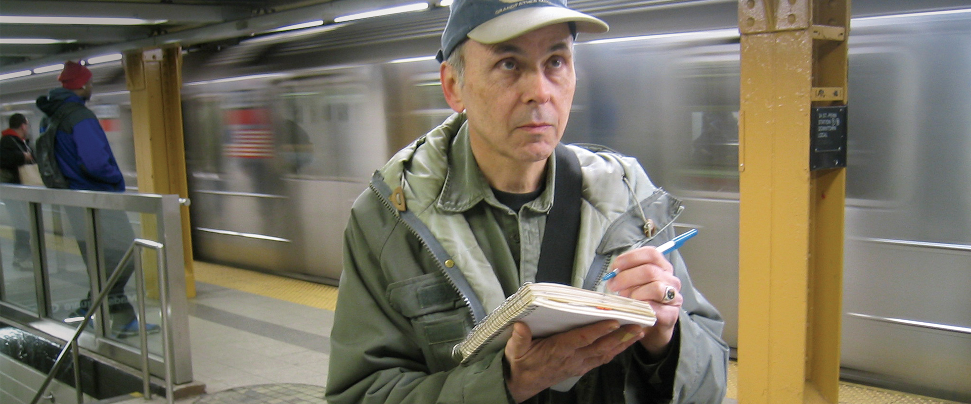 A man with a notebook stands on a platform and considers a train in motion.