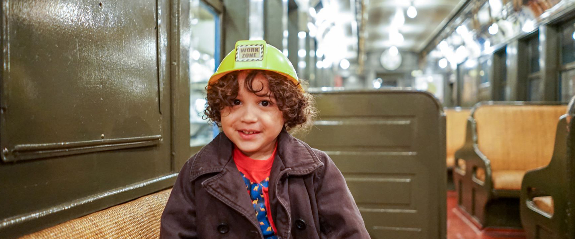 Young child smiles on old Elevated Train wearing yellow construction hat.