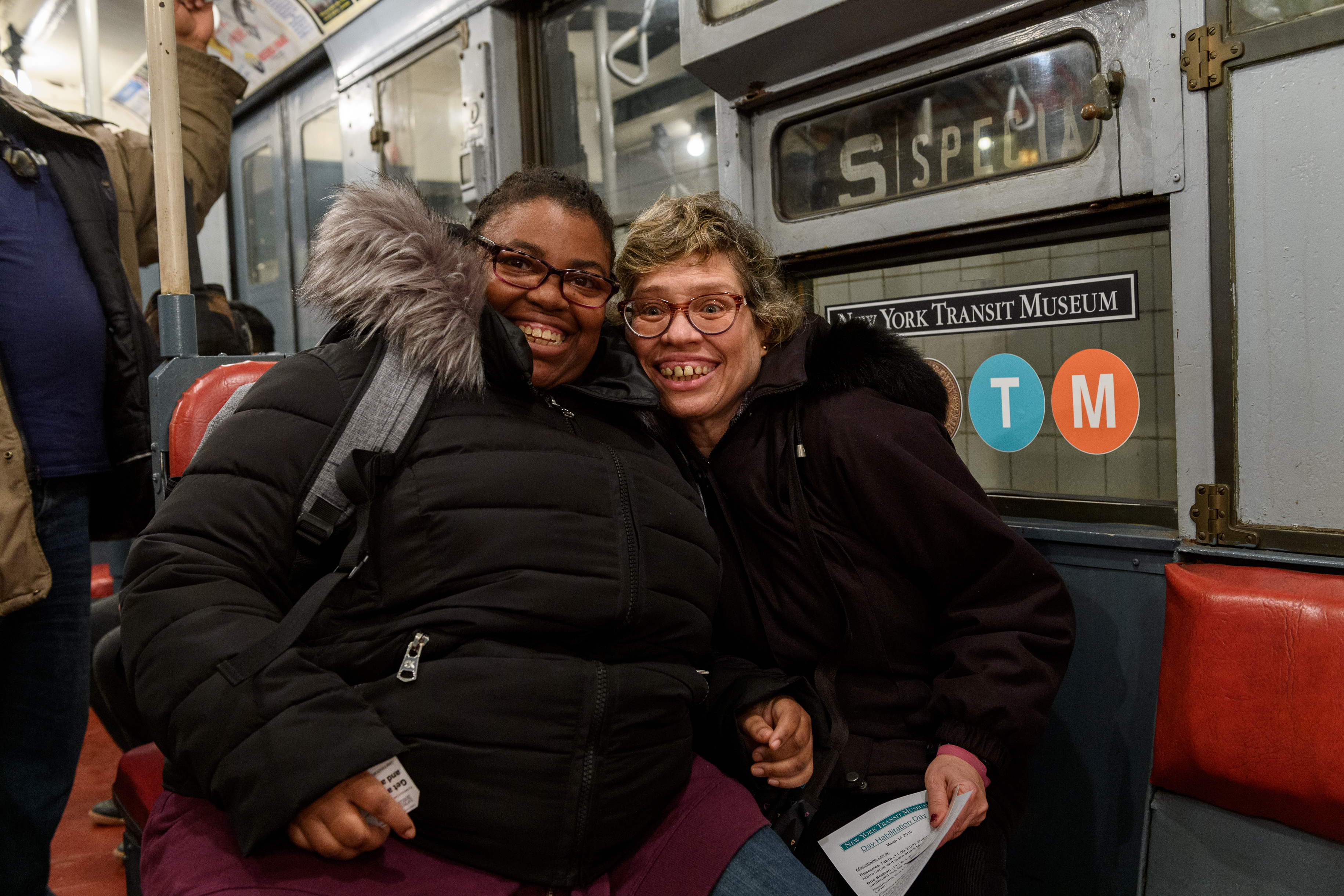 Two women sit close together with big smiles on a subway car.