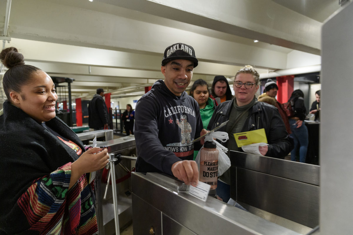 A man swipes a MetroCard at a turnstile with two women watching on either side of him.