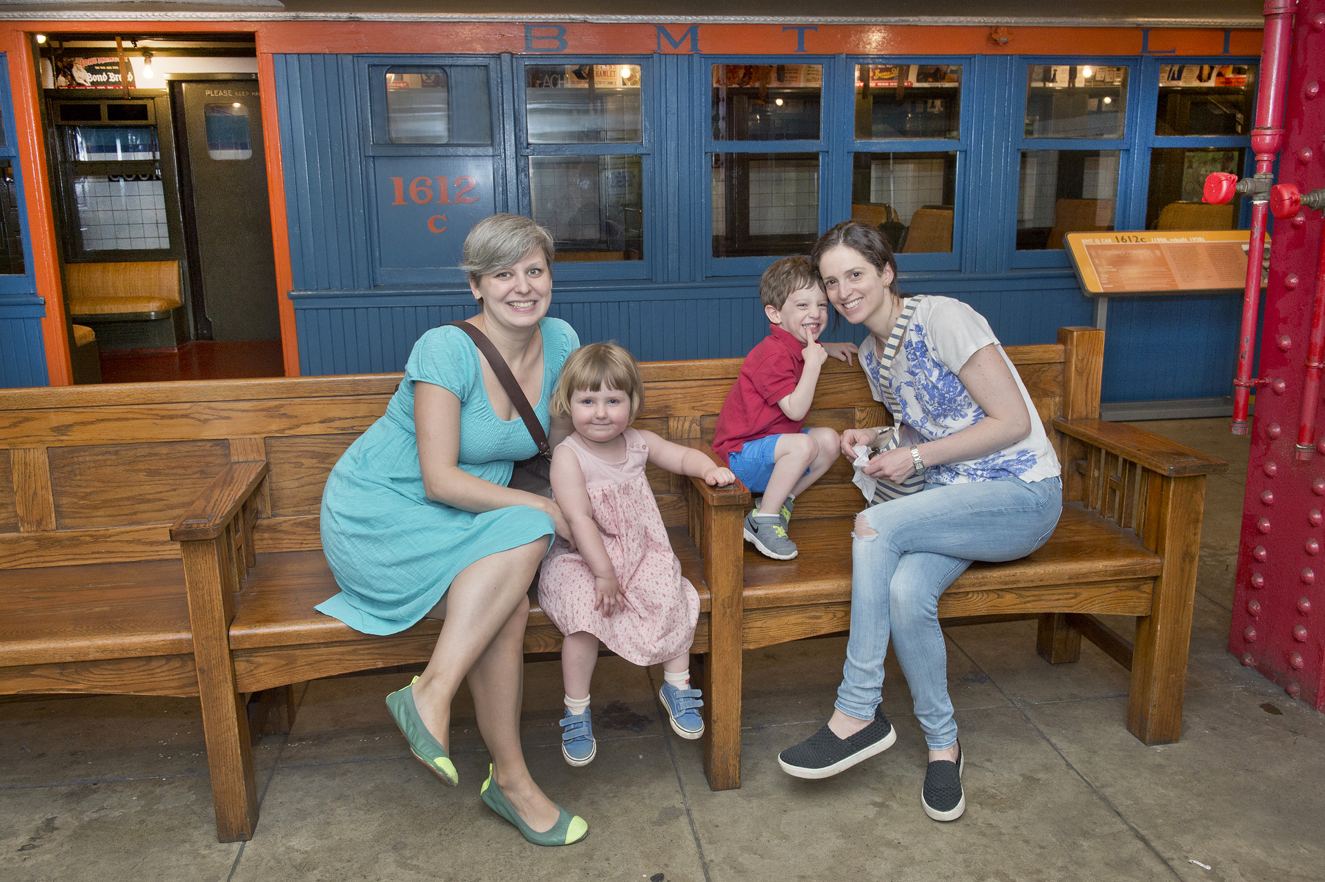 A young girl and boy sit together with two women in front of a blue and orange vintage train car.