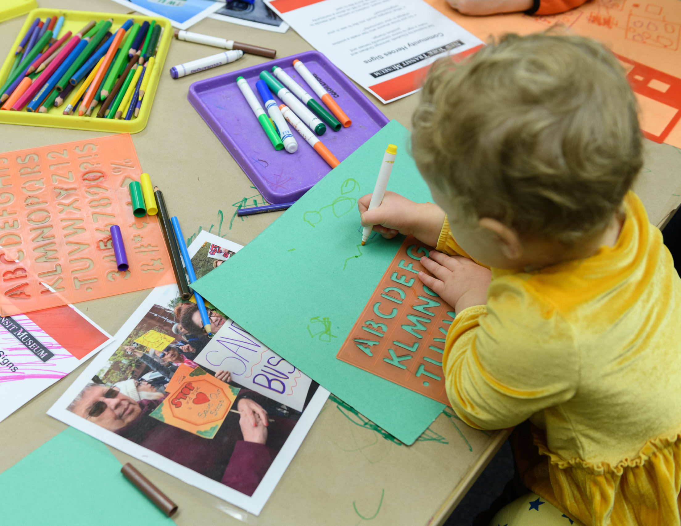 A young child surrounded by art supplies draws on paper with a marker.