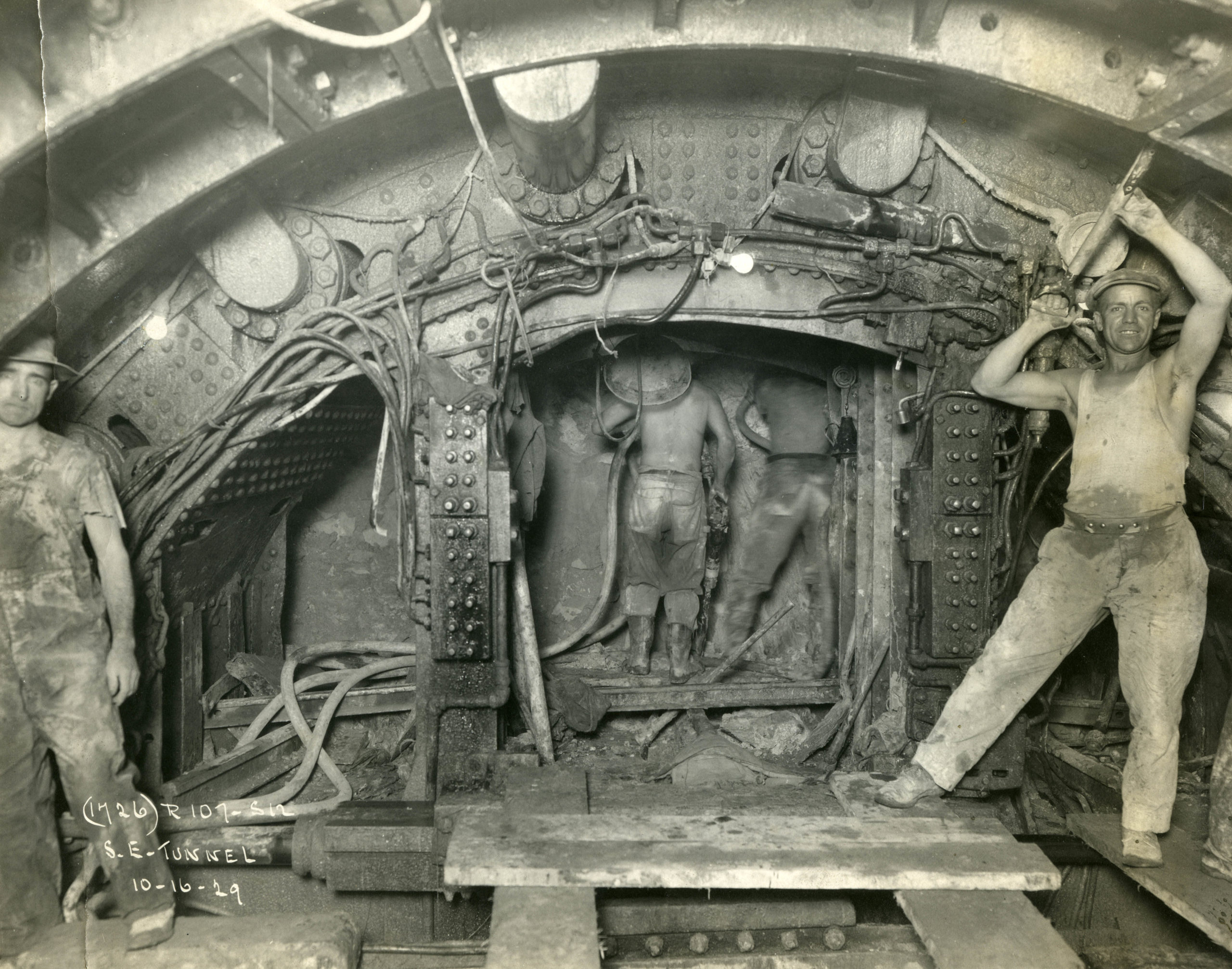 Black and white photo of four tunnel workers. Two are at the far edge of the image, facing the camera. Two are standing in the middle background, with their backs towards the camera.