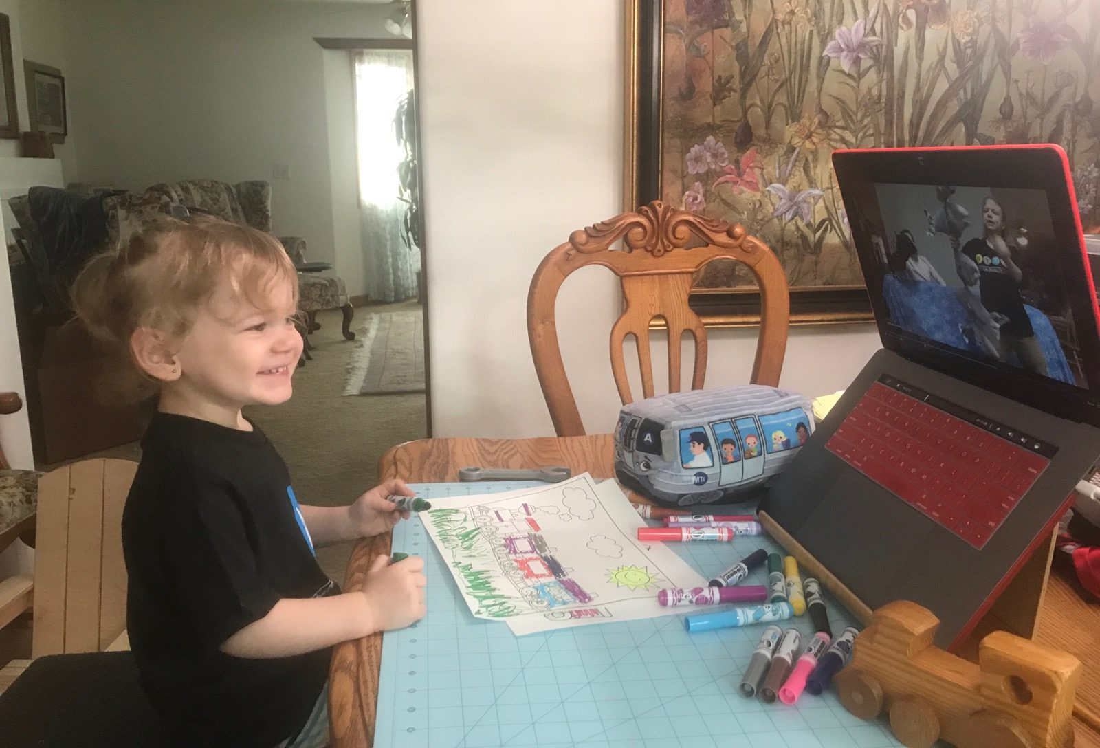 Child makes art at a table at home and smiles at a Museum educator on a computer screen.