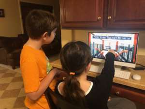 A boy and girl engage during a virtual program. Their computer screen shows the New York Transit Museum logo and a photo of inside the Museum.