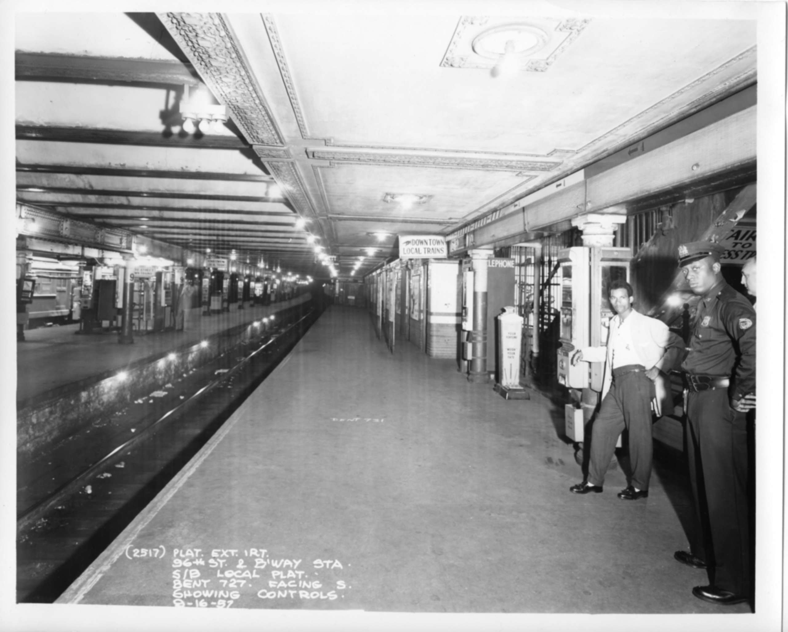 A black and white photograph in an underground subway station that looks down the subway tracks and platform. Two men stand on the far right looking at the camera.