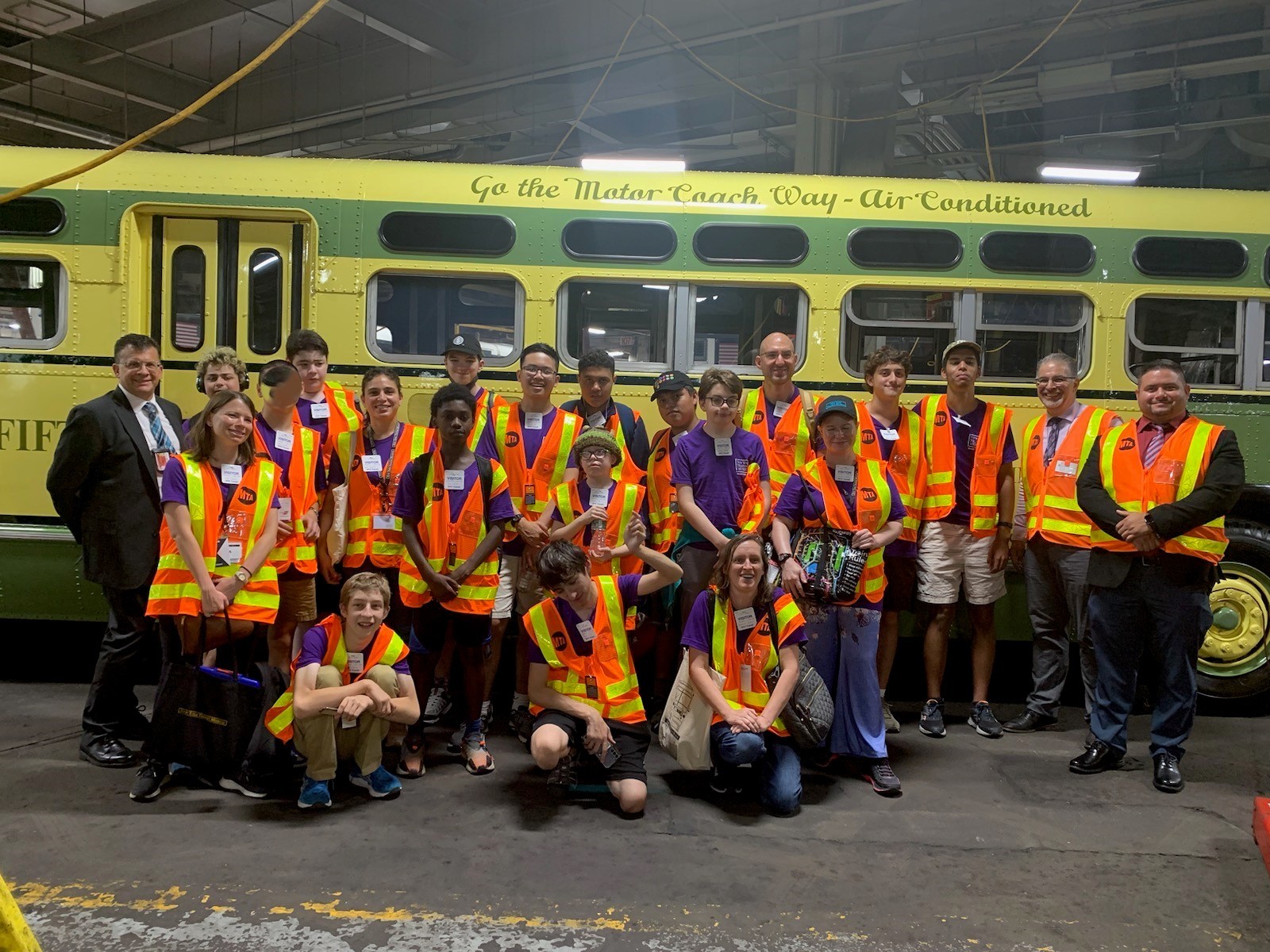 A large group of teens and adults wear orange safety vests standing in front of a vintage bus.