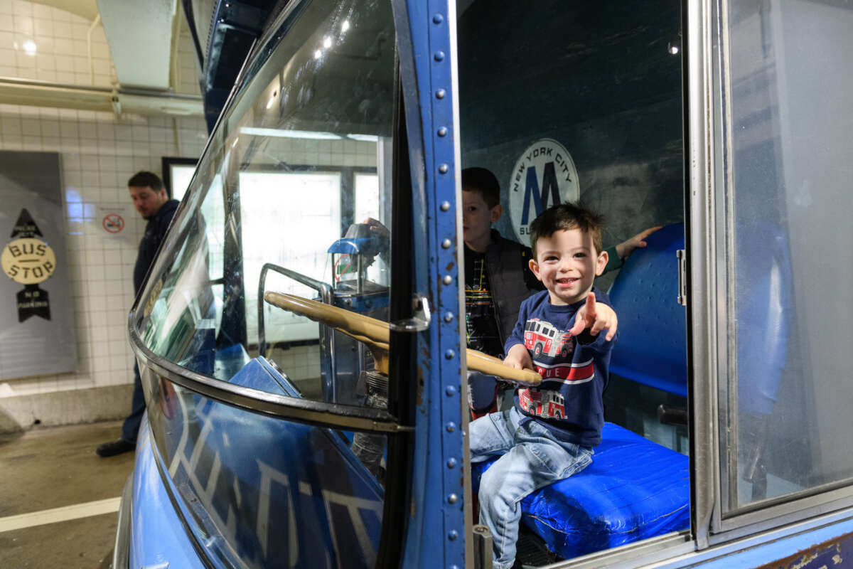 Child sitting in driver's seat of bus, pointing at camera