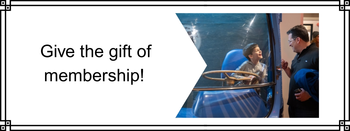 Banner image with a white background and black lines around the boarder. A text box on the left says “Give the gift of membership!”. On the right, there is a photo of a young boy sitting in the driver’s seat of a bus. The picture is taken through the windshield, the seats and steering wheel are blue. The young boy in the driver’s seat is smiling to his father, who is standing to the right of the bus, smiling back through the driver’s side window. 