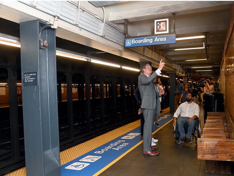 Two men on a subway platform, with one man standing pointing to wheelchair signage and another man in a wheelchair.