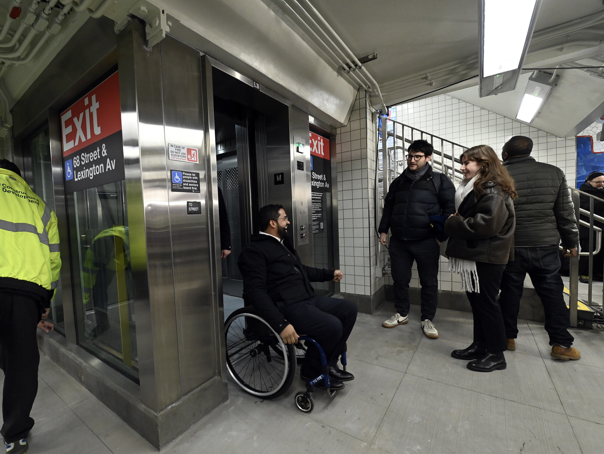 Man in wheelchair exits elevator onto subway mezzanine.
