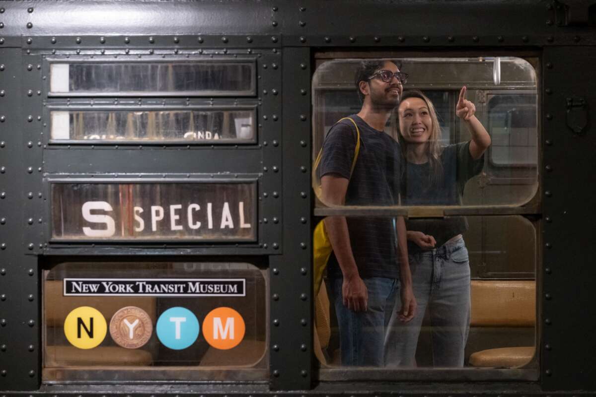 Two visitors in the window of a vintage train car, woman seen pointing at something inside, both are smiling. Train roll sign reads S Special and there is a New York Transit Museum logo with NYTM graphic on the window.