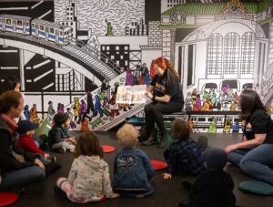 A NY Transit Museum staff member reads a children's book to toddlers and their care givers in front of a colorful mural.