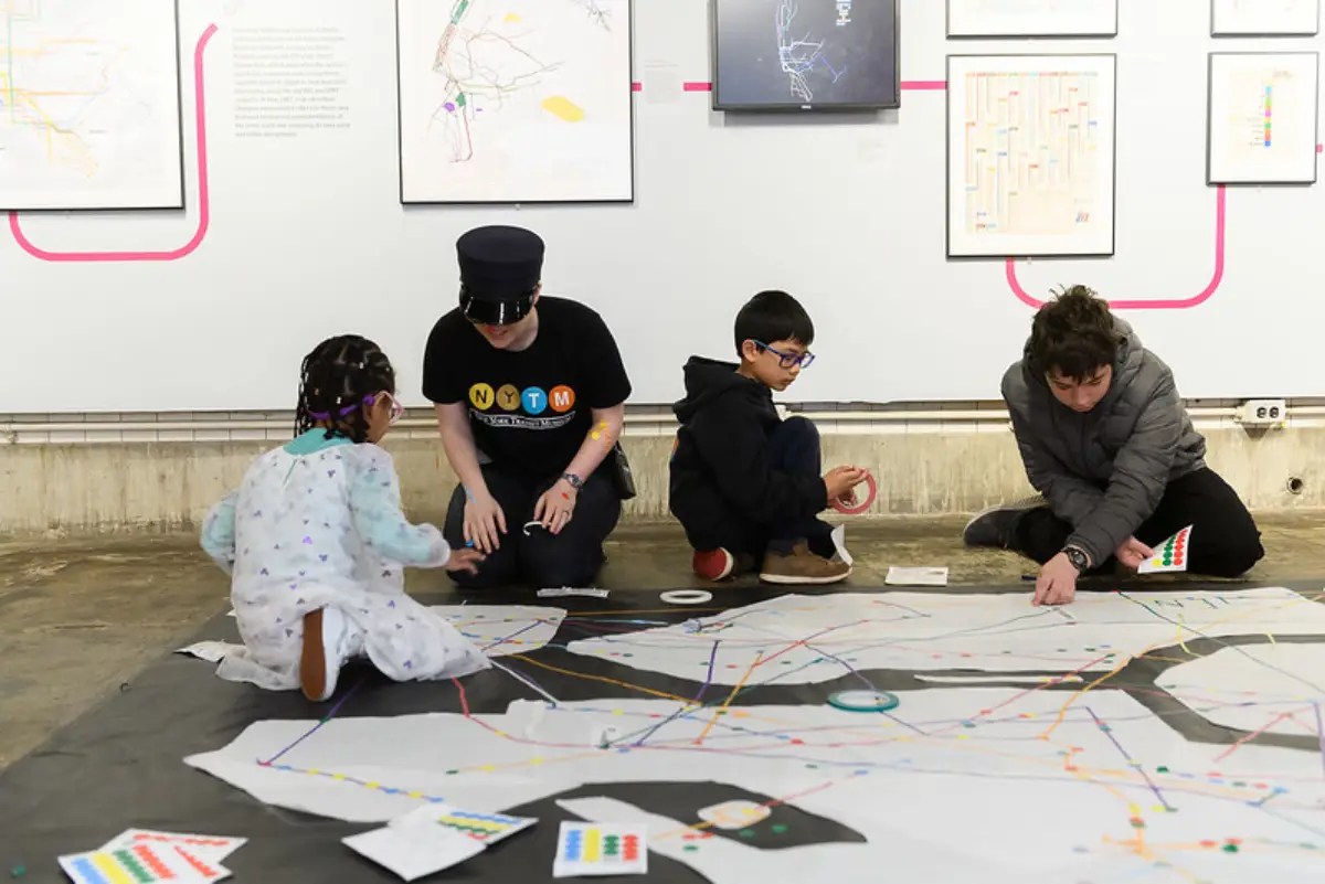 Museum staff member wearing conductor's hat sits with children participating in an art activity