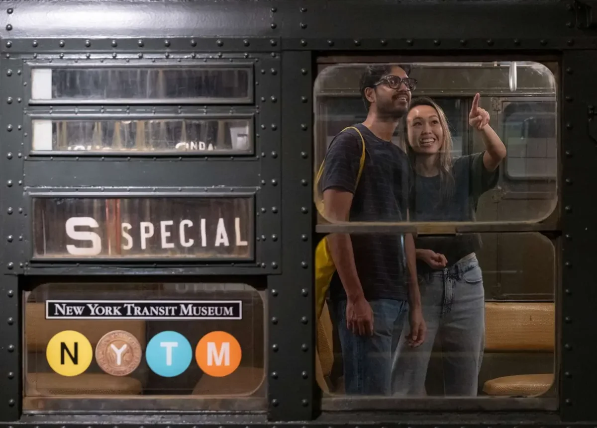Seen through the window, two visitors explore a vintage train. Woman is pointing up at vintage ads.