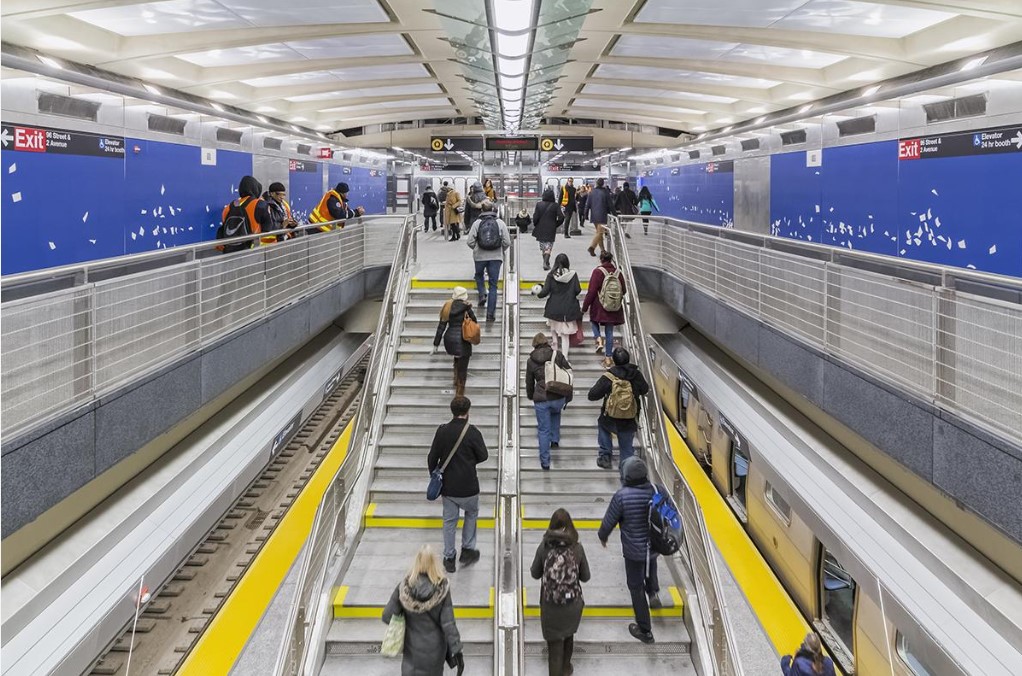 People walk upstairs from subway platform to wide mezzanine with blue artwork on walls.