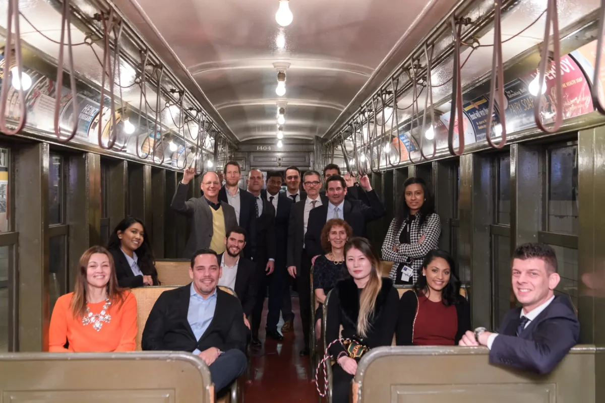 Men and women from the same company pose in a restored 1904 Brooklyn Union elevated car with lightbulbs and straps.