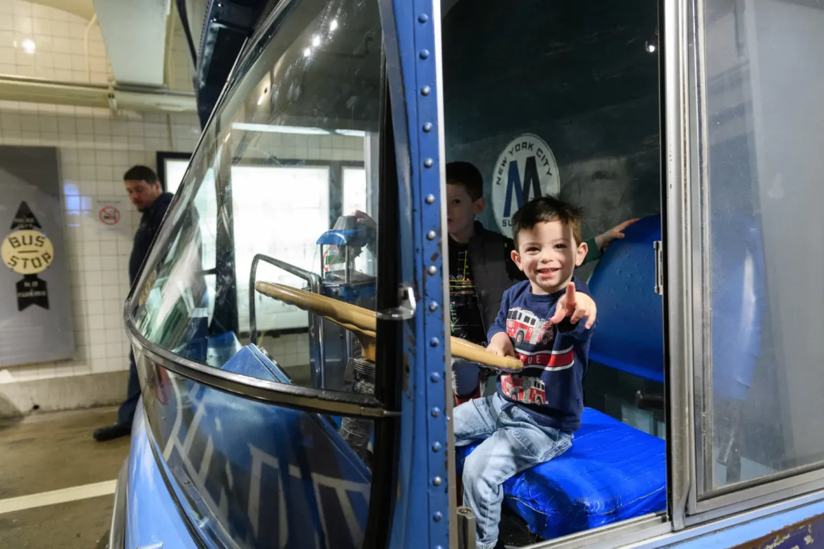 An adorable toddler boy points and pretends to drive a New York City bus.