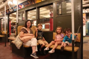 A mom sits on a vintage train at the New York Transit Museum with her three happy kids.