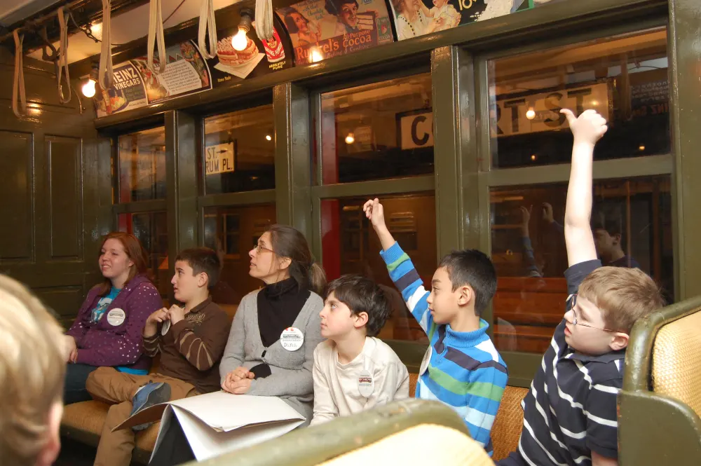 Group of students sitting in vintage subway car excitedly raising their hands to answer questions