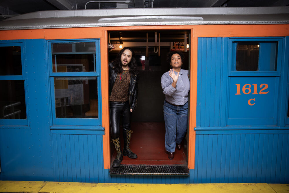 A man and women stand in vintage subway car, singing.