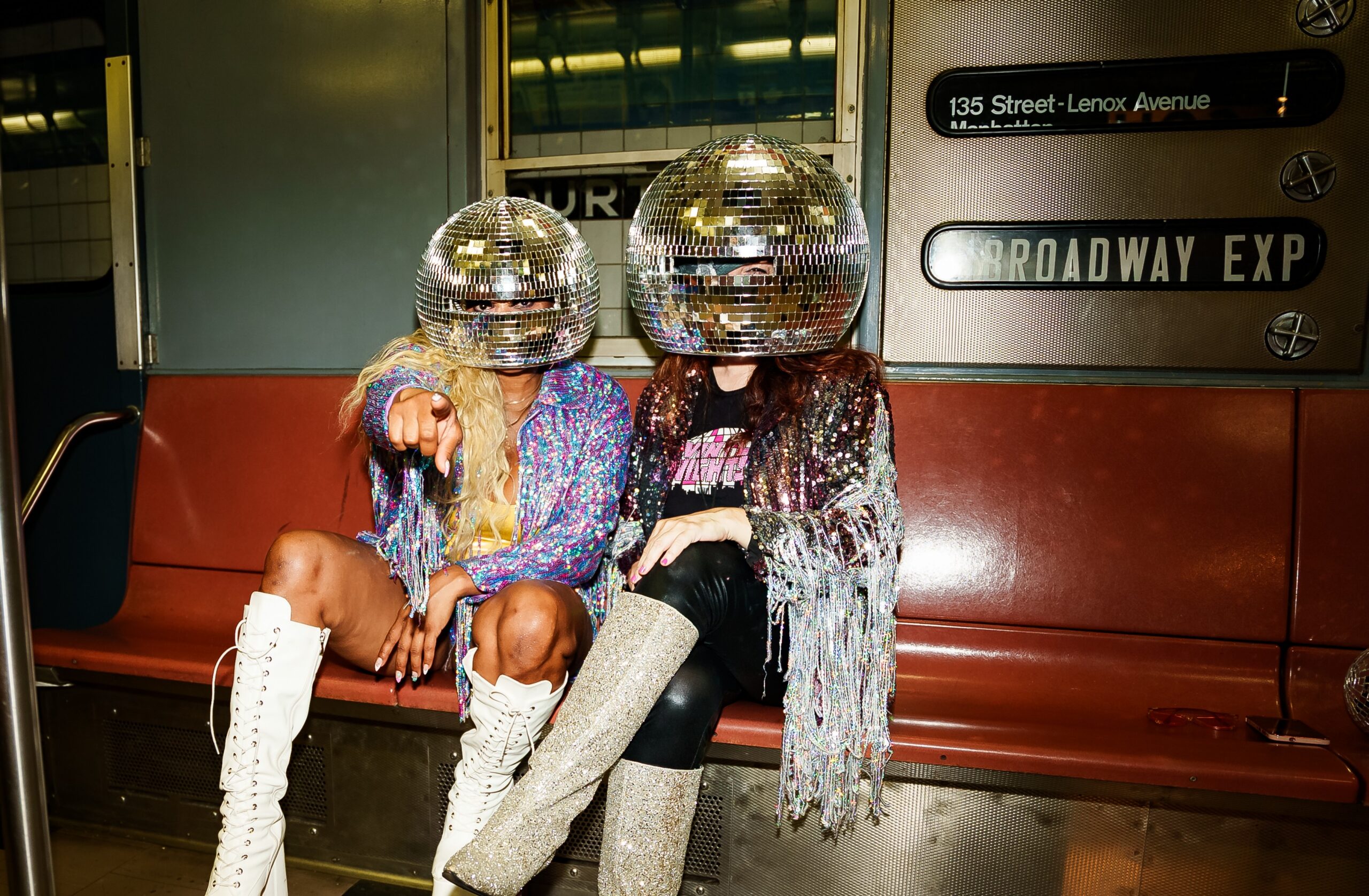 Two woman in sparkly clothing and a mirror ball helmet sit inside a subway car.