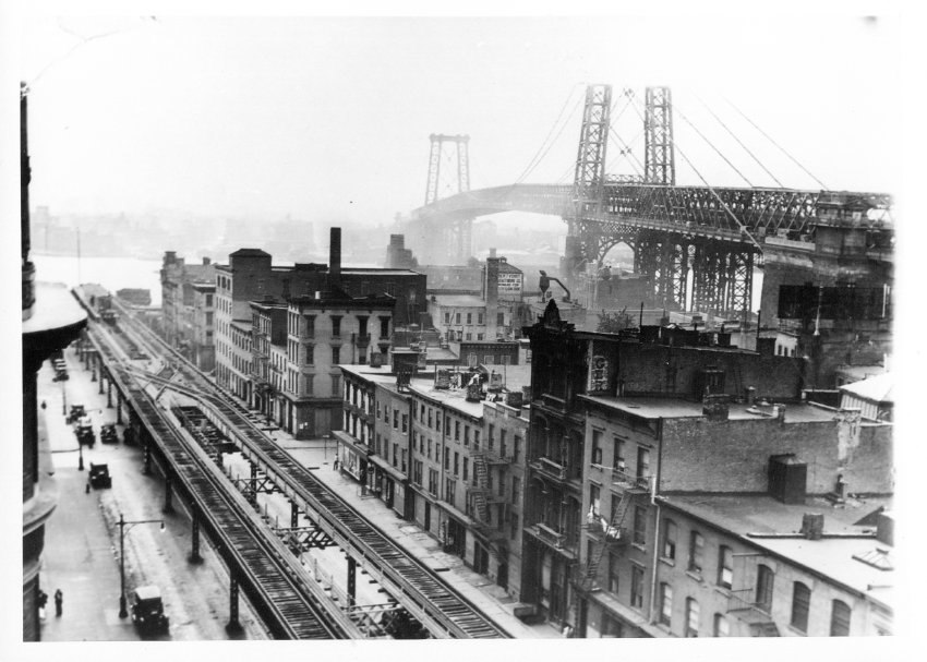 Elevated tracks run between buildings. The Williamsburg Bridge can be seen in the near distance.