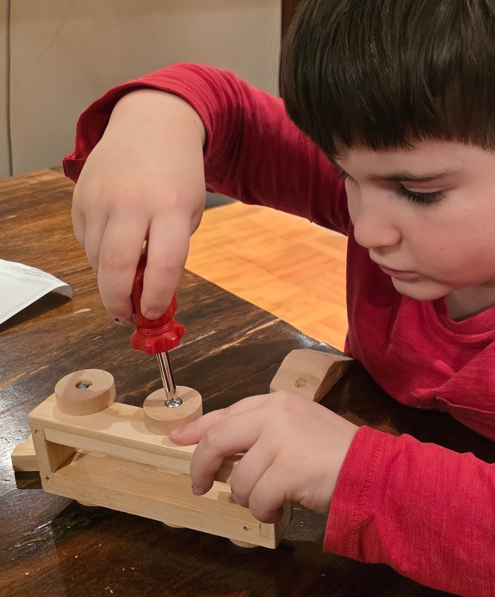 Child wearing a red shirt using a screwdriver to tighten a wheel on a wooden model train.