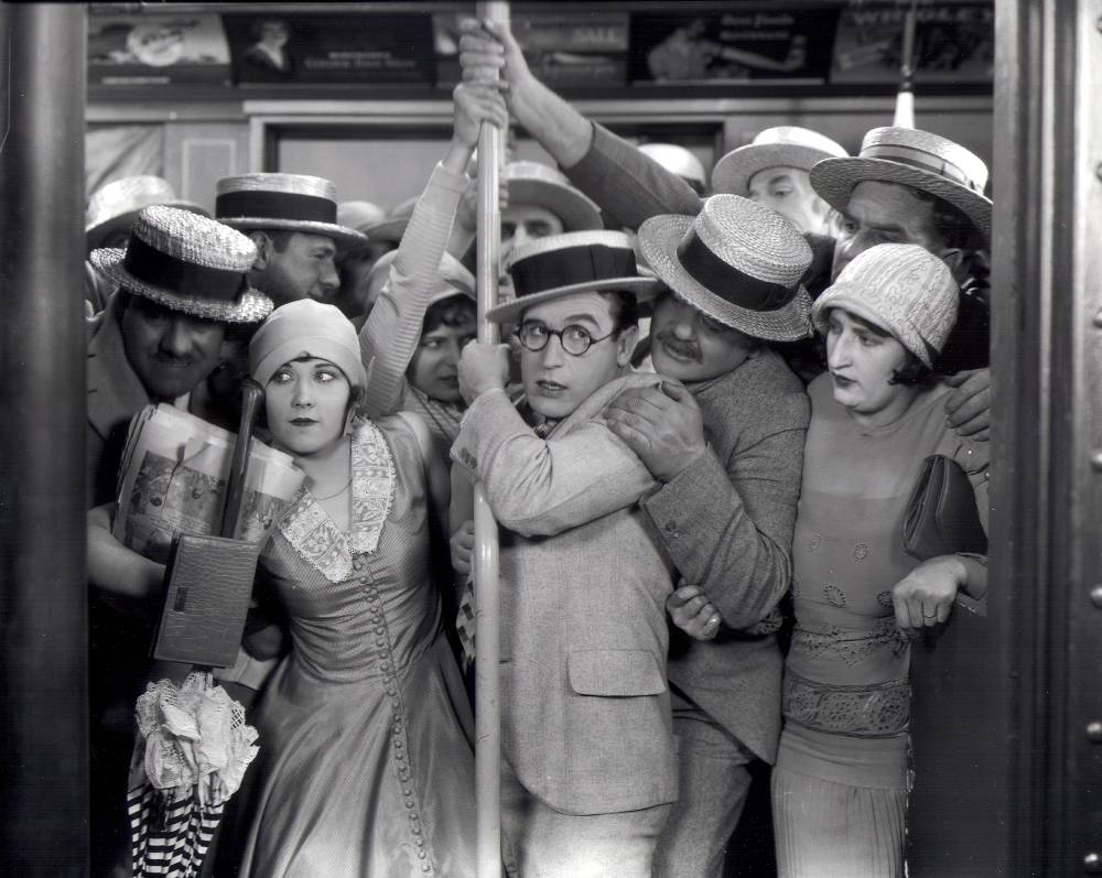 A black and white photograph show men and women crowded in a subway car holding onto a pole.