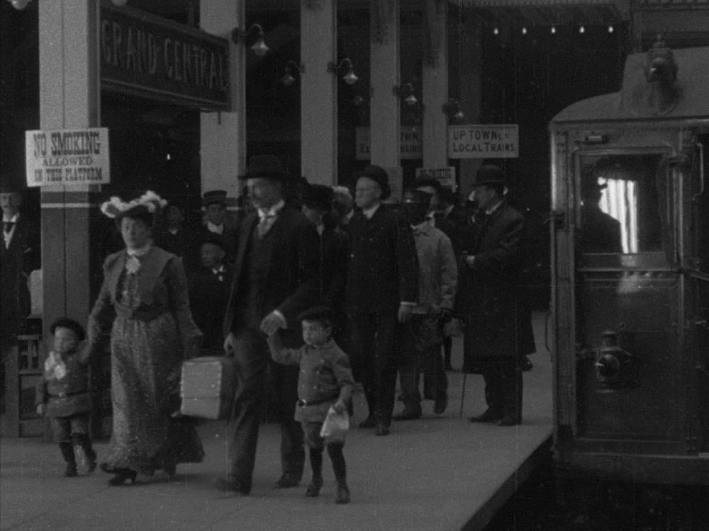 In a black and white photo, men, women, and children are on a platform next to a train.