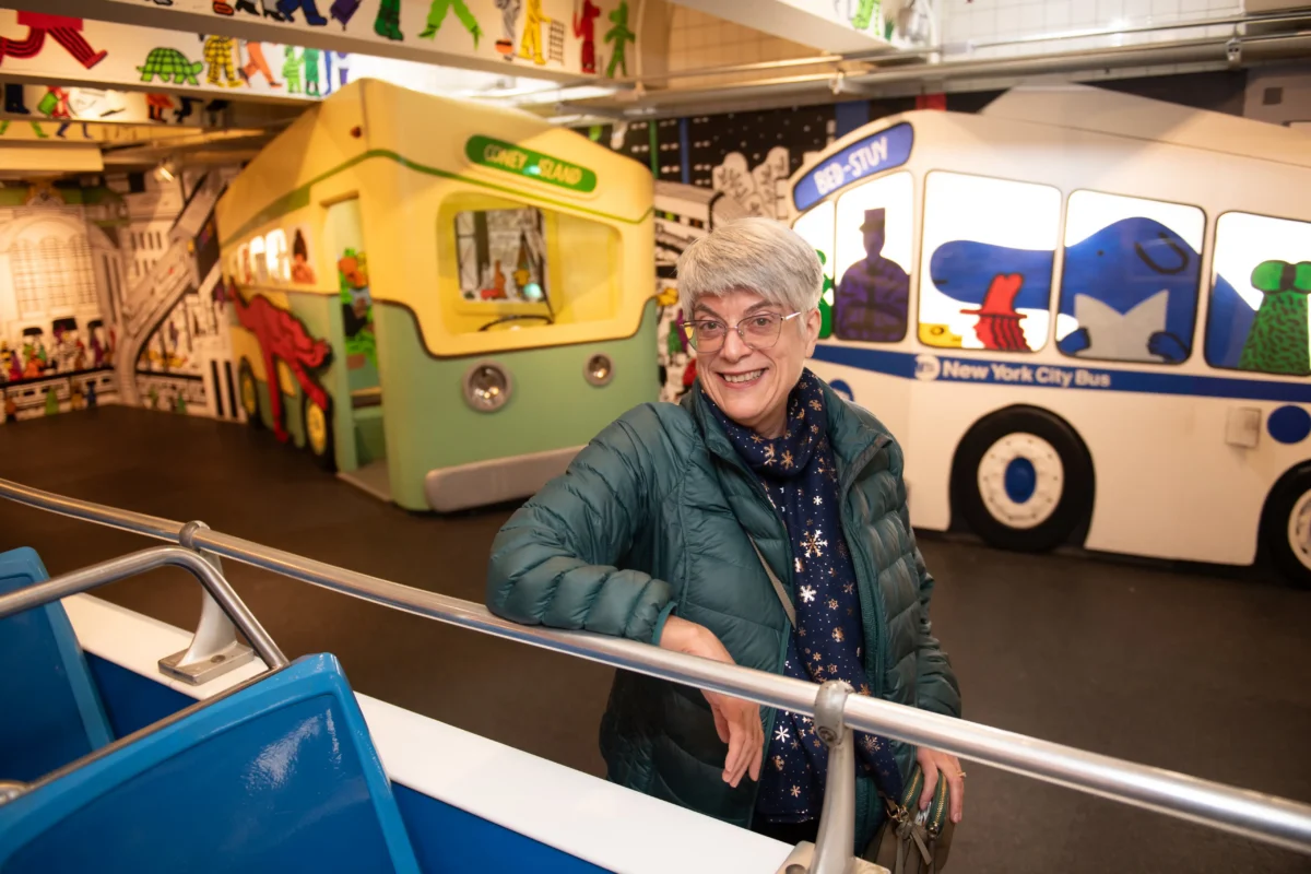 A woman smiles with several buses in the background at the New York Transit Museum.